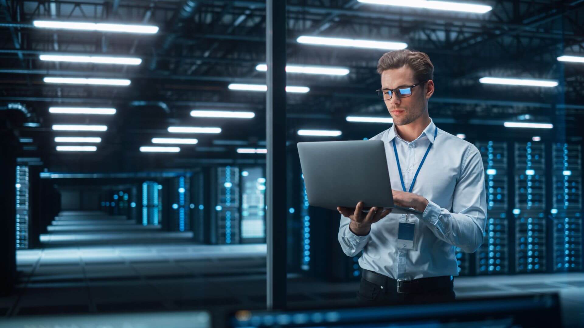 A Man Wearing Glasses, an IT Specialist, Holds a Laptop in a Server Room, Managing Technical Tasks