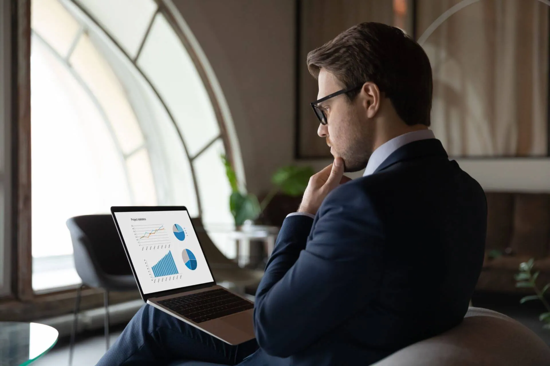 IT Expert in a Suit Analyzing It Solutions on a Laptop in a Modern Office Lounge